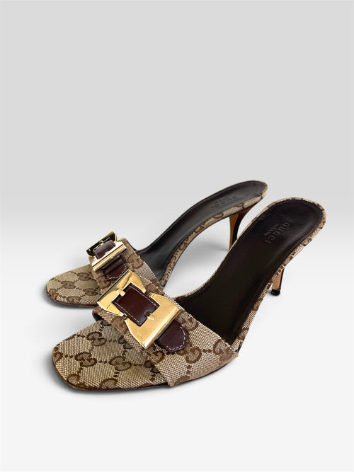 A pair of Gucci GG Supreme monogram mule sandals with gold-tone and dark brown buckle details, shown on a light gray background.
