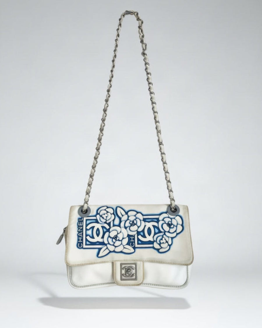 A Chanel shoulder bag in off-white leather with a blue and white floral camellia pattern on the front flap, featuring a silver-tone chain strap and a silver CC turn-lock closure.