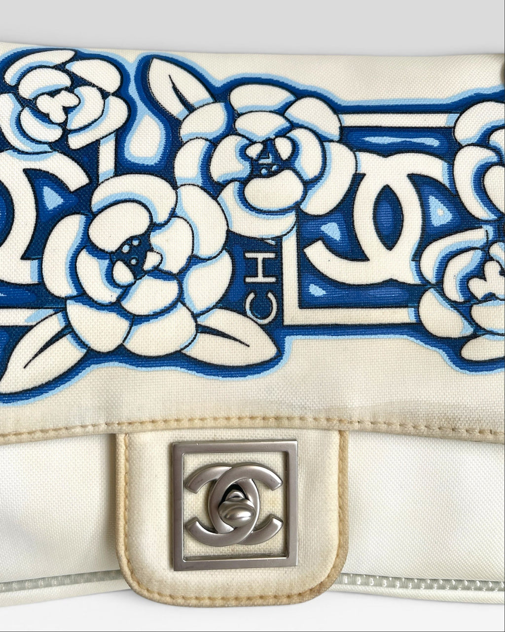Close-up of a Chanel shoulder bag flap featuring blue and white embroidered floral and interlocking CC motifs, with a silver-tone interlocking CC turn-lock closure on a light beige fabric base.